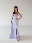 A woman modeling a strapless lavender satin maxi gown with a thigh-high slit, standing against a plain light gray studio background. / Straight neckline A-line gown with back bow detail.