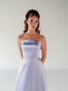 A woman modeling a strapless light lavender satin dress against a plain light gray studio background. / Straight neckline A-line gown with back bow detail.