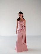 A young woman modeling a strapless dusty pink satin maxi dress with a draped waist and side bow, set against a plain light gray studio background. / Slim column gown with draped bodice and bow detail.
