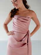 A woman wearing a strapless dusty pink satin dress with draped ruching and a side tie detail, photographed against a plain light gray studio background. / Slim column gown with draped bodice and bow detail.