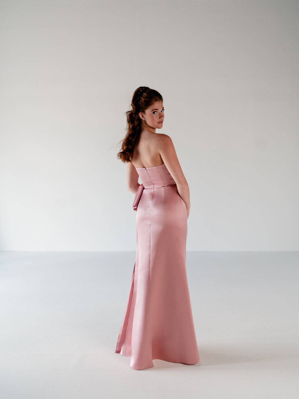 A woman modeling a strapless dusty pink satin gown with a matching waist bow, photographed from the back in a minimalist studio setting. / Slim column gown with draped bodice and bow detail.