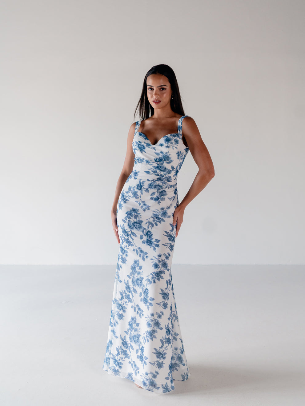 A woman modeling a sleeveless white maxi dress with a blue floral print and thin shoulder straps, standing against a plain light gray studio background. / Slim chiffon gown with bodice and skirt pleats and a cowl back.