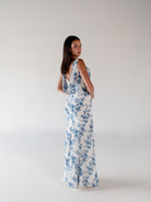 A woman modeling a sleeveless white maxi dress with a blue floral print, shown from the back against a plain light gray studio background. / Slim chiffon gown with bodice and skirt pleats and a cowl back.
