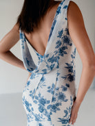 A woman wearing a sleeveless white dress with a blue floral print, shown from the back with a deep open back and draped neckline. / Slim chiffon gown with bodice and skirt pleats and a cowl back.