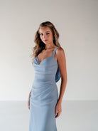 A woman modeling a light blue sleeveless wrap-style dress with thin straps and a fitted silhouette, standing against a plain light gray studio background. / Slim chiffon gown with bodice and skirt pleats and a cowl back.