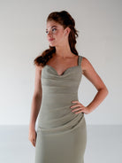 A woman modeling a sleeveless, cowl-neck, bodycon dress in a muted sage green color against a plain light gray studio background. / Slim chiffon gown with bodice and skirt pleats and a cowl back.