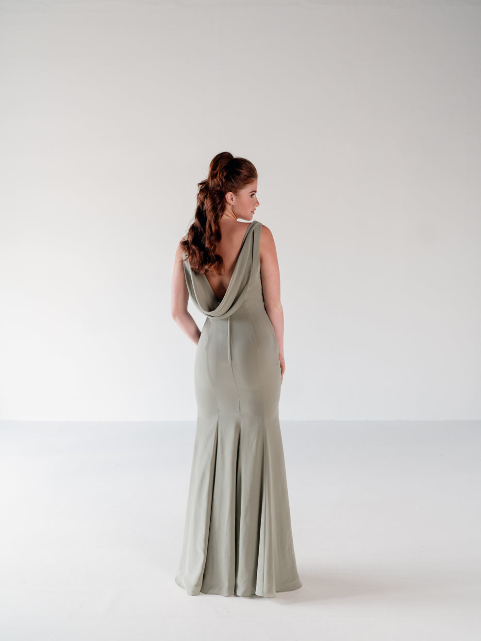 A woman wearing a floor-length sage green gown, photographed from the back in a studio setting with a plain light background. / Slim chiffon gown with bodice and skirt pleats and a cowl back.