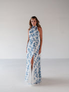 A woman modeling a sleeveless halter-neck maxi dress with a high thigh slit, featuring a blue floral print on a white background, set against a plain light gray studio backdrop. / Draped halter gown with A-line skirt.