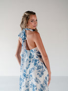 A woman modeling a white halter dress with a blue floral print, shown from the back and side against a plain light gray studio background. / Draped halter gown with A-line skirt.