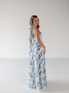 A woman modeling a sleeveless white maxi dress with a blue floral print and tie-shoulder back detail, photographed in a minimalist studio setting. / Draped halter gown with A-line skirt.