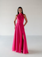 A woman modeling a bright hot pink halter-neck floor-length gown with a high thigh slit, standing against a plain light gray studio background. / Draped halter gown with A-line skirt.