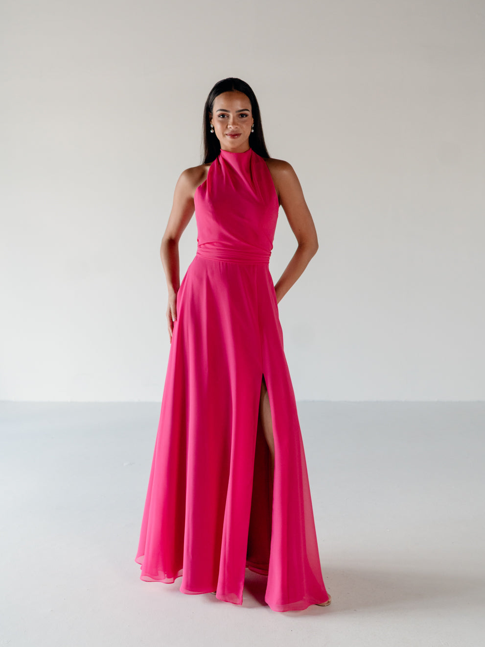 A woman modeling a bright hot pink halter-neck floor-length gown with a high thigh slit, standing against a plain light gray studio background. / Draped halter gown with A-line skirt.