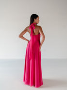 A woman modeling a bright pink halter-neck maxi dress from the back, featuring a long draped tie at the back of the neck and a flowing satin-like skirt, set against a plain light gray studio background. / Draped halter gown with A-line skirt.