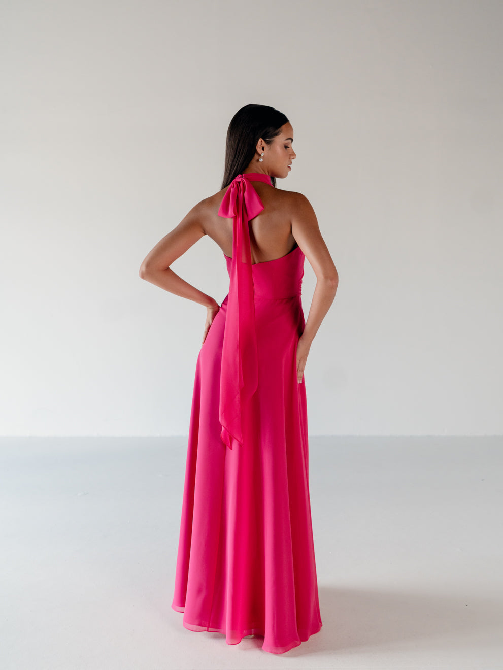 A woman modeling a bright pink halter-neck maxi dress from the back, featuring a long draped tie at the back of the neck and a flowing satin-like skirt, set against a plain light gray studio background. / Draped halter gown with A-line skirt.