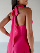 A woman shown from the back wearing a bright fuchsia halter dress with a long draped tie detail over one shoulder, photographed against a clean light gray studio background. / Draped halter gown with A-line skirt.