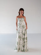 A woman modeling a strapless white maxi dress with a green leaf pattern, standing against a plain light gray studio background. / Basque waist draped bodice with an A-line skirt.