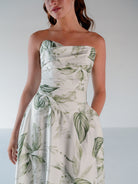 A woman wearing a strapless white dress with a green tropical leaf print, shown from the shoulders down against a plain light gray studio background. / Basque waist draped bodice with an A-line skirt.