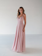 A woman modeling a floor-length, sleeveless dusty pink gown with thin spaghetti straps and a V-neckline, standing against a plain light gray studio background. / A-line gown with double princess seam detail.