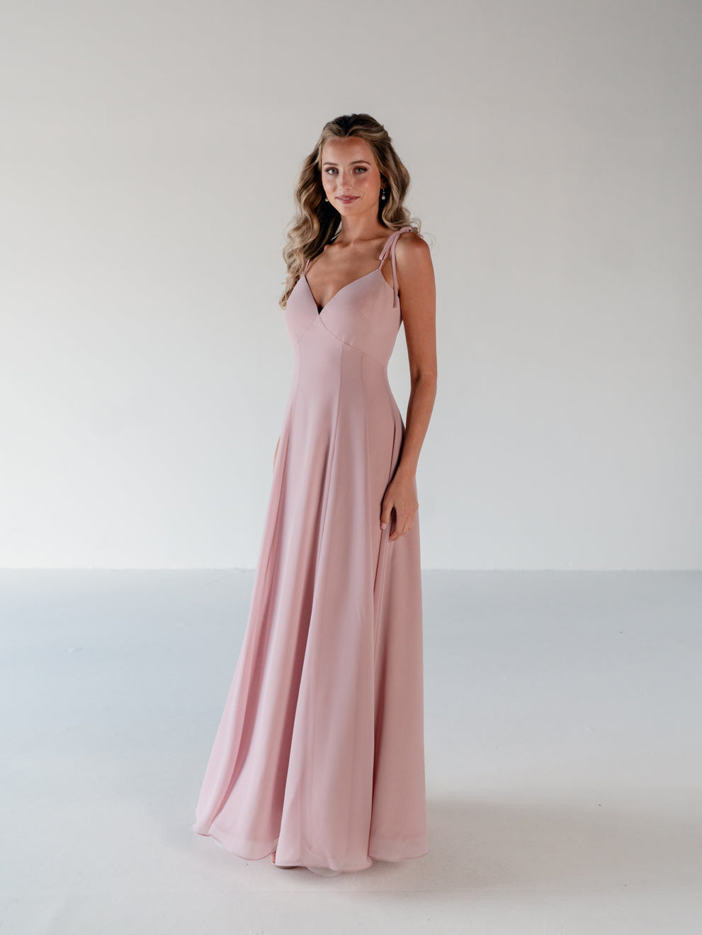 A woman modeling a floor-length, sleeveless dusty pink gown with thin spaghetti straps and a V-neckline, standing against a plain light gray studio background. / A-line gown with double princess seam detail.