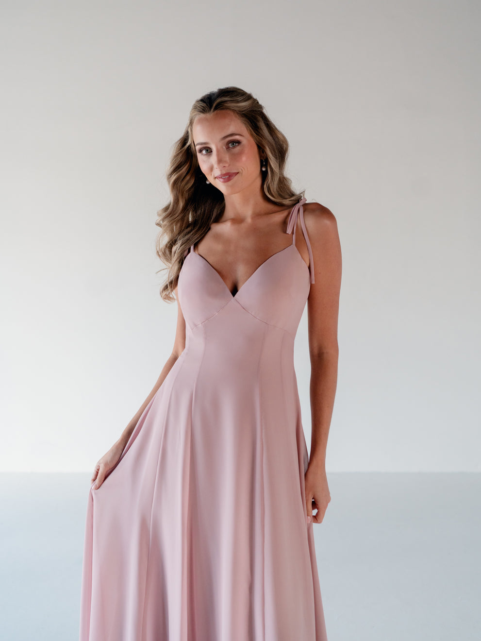 A woman modeling a floor-length dusty pink gown with thin shoulder straps, a V-neckline, and a flowing skirt against a plain light gray studio background. / A-line gown with double princess seam detail.