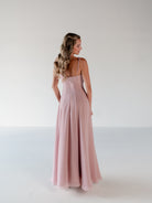 A woman modeling a floor-length dusty pink sleeveless dress, shown from the back in a studio setting with a light gray background. / A-line gown with double princess seam detail.