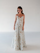 A woman modeling a floor-length sleeveless gown with thin straps and a soft floral print in pale blush, beige, and muted green on a light gray background. / A-line gown with double princess seam detail.