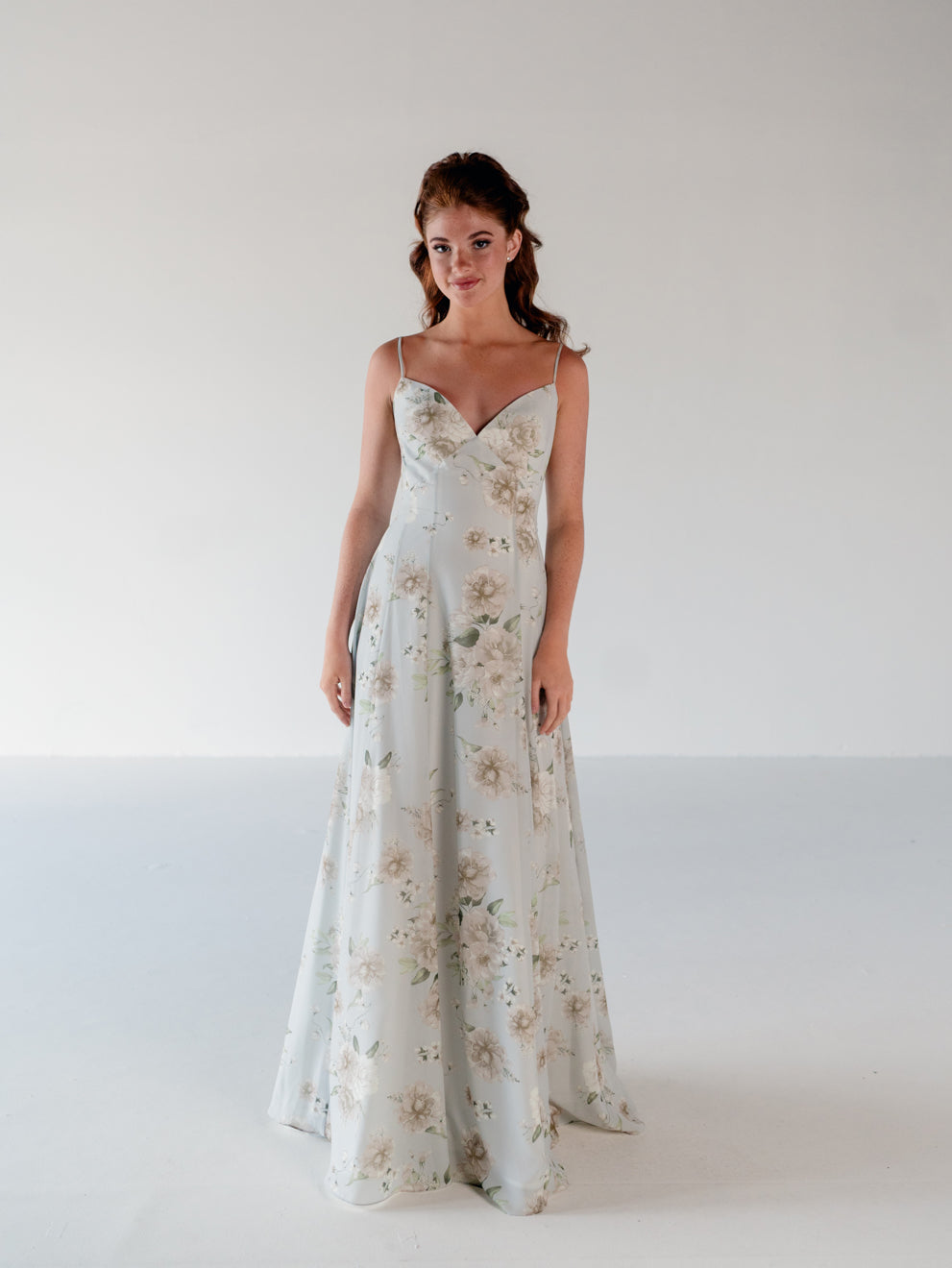 A woman modeling a floor-length sleeveless gown with thin straps and a soft floral print in pale blush, beige, and muted green on a light gray background. / A-line gown with double princess seam detail.