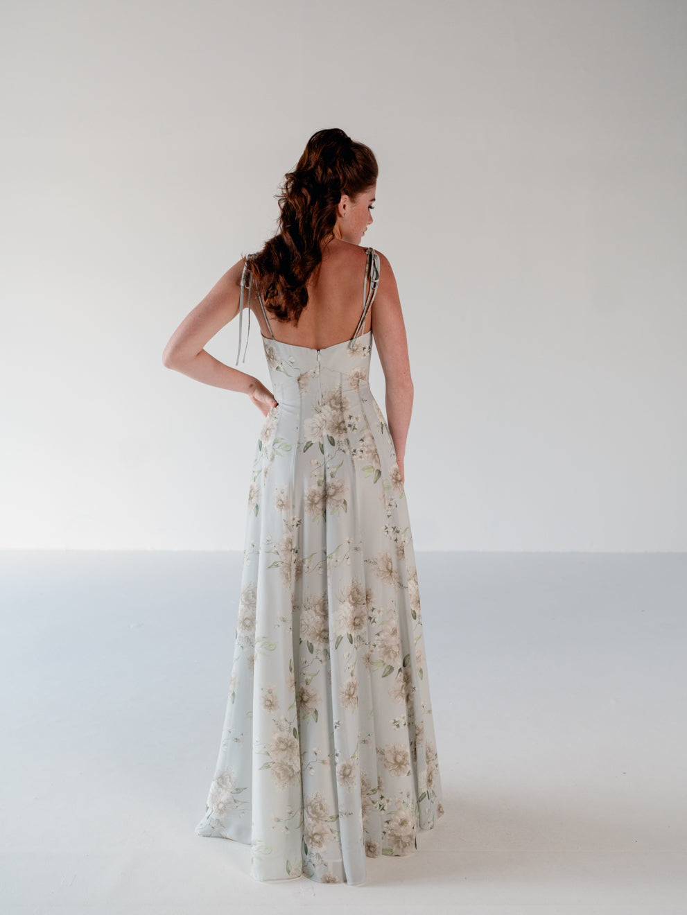 A woman stands with her back to the camera wearing a floor-length sleeveless dress with thin straps and a soft floral print in pale blue, beige, and cream against a plain light gray studio background. / A-line gown with double princess seam detail.