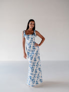 A woman modeling a sleeveless white maxi dress with a blue floral print and tie-shoulder straps, standing against a plain light gray studio background. / Slim satin gown with soft square neckline and bow detail at the shoulders.