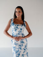 A woman modeling a sleeveless white dress with a blue floral toile pattern, featuring a square neckline and wide shoulder straps, set against a plain light gray background. / Slim satin gown with soft square neckline and bow detail at the shoulders.