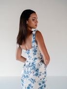 A woman wearing a sleeveless white dress with a blue floral print, shown from the back and side against a plain light gray studio background. / Slim satin gown with soft square neckline and bow detail at the shoulders.