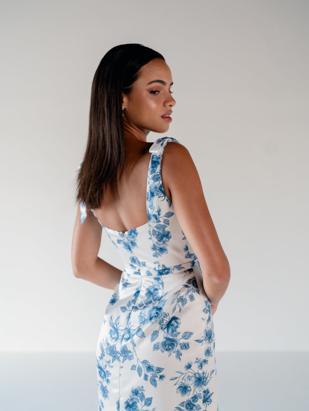 A woman wearing a sleeveless white dress with a blue floral print, shown from the back and side against a plain light gray studio background. / Slim satin gown with soft square neckline and bow detail at the shoulders.