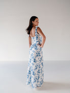 A woman modeling a sleeveless white maxi dress with a blue floral print, shown from the back in a studio setting with a plain light background. / Slim satin gown with soft square neckline and bow detail at the shoulders.
