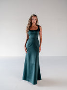 A woman modeling a floor-length teal green satin gown with black ribbon shoulder straps, standing against a neutral light gray studio background. / Slim satin gown with soft square neckline and bow detail at the shoulders.