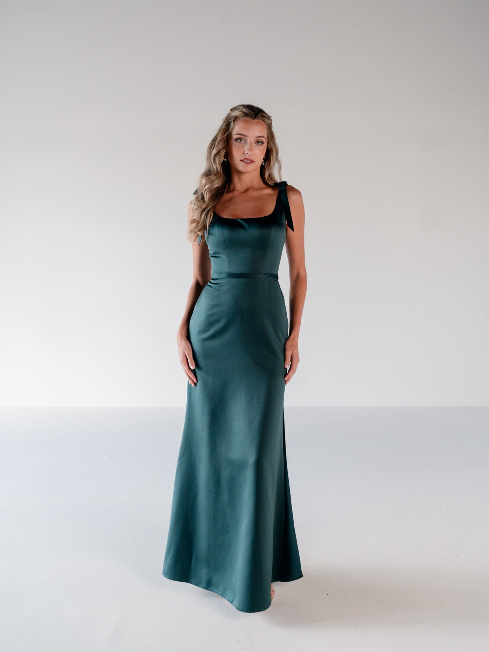 A woman modeling a floor-length teal green satin gown with black ribbon shoulder straps, standing against a neutral light gray studio background. / Slim satin gown with soft square neckline and bow detail at the shoulders.