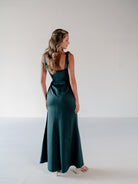 A woman wearing a dark teal green satin maxi dress, shown from the back in a studio setting with a white background. / Slim satin gown with soft square neckline and bow detail at the shoulders.