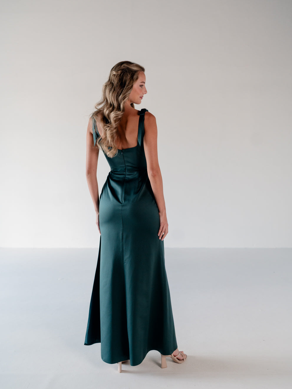 A woman wearing a dark teal green satin maxi dress, shown from the back in a studio setting with a white background. / Slim satin gown with soft square neckline and bow detail at the shoulders.