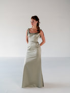 A woman modeling a floor-length sage green satin gown with tie straps at the shoulders, standing against a plain light gray studio background. / Slim satin gown with soft square neckline and bow detail at the shoulders.