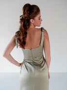 A woman with long wavy brown hair is shown from the back wearing a sleeveless, structured dress in a light sage green or olive-gray satin-like fabric. The dress has wide shoulder straps with bow details and a gathered waist seam. / Slim satin gown with soft square neckline and bow detail at the shoulders.