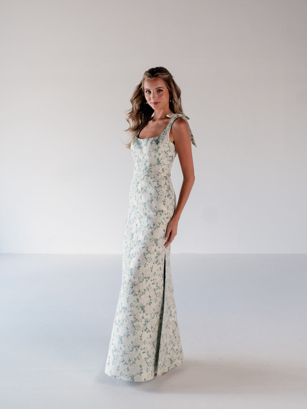 A woman modeling a sleeveless, floor-length floral dress with a fitted silhouette and a front slit, standing against a plain light gray studio background. / Slim gown with soft square neckline and bow detail at the shoulders.