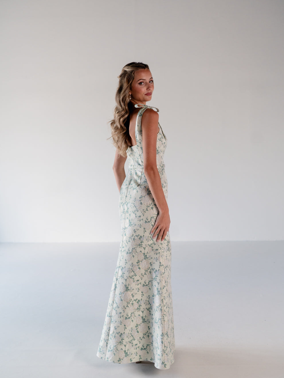 A woman modeling a sleeveless, floor-length floral dress with a tie-neck detail, standing in profile against a plain light gray studio background. / Slim gown with soft square neckline and bow detail at the shoulders.