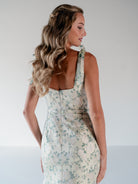 A woman shown from the back wearing a sleeveless, scoop-neck dress with a light beige base and a delicate blue and green floral pattern. The dress features a structured bodice, a defined waist, and a fitted skirt. / Slim gown with soft square neckline and bow detail at the shoulders.