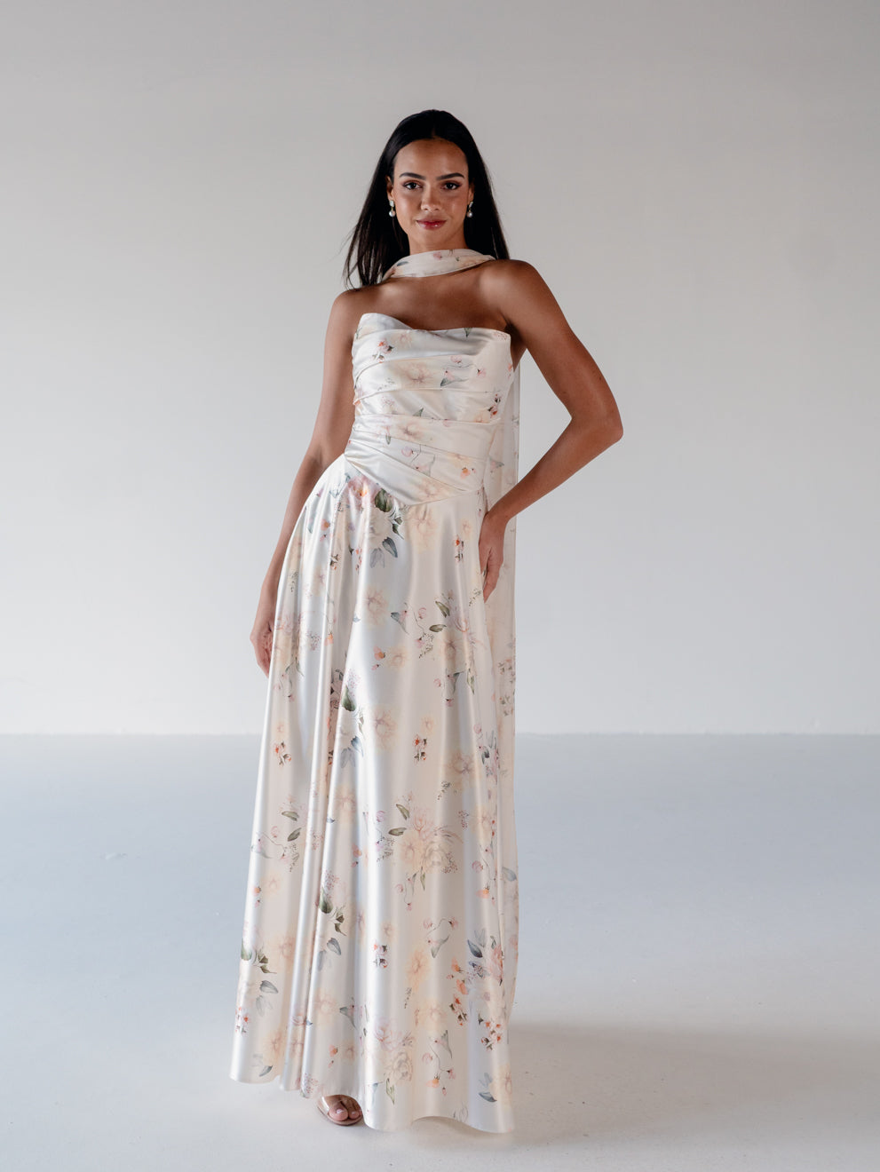 A woman modeling a strapless, floor-length satin gown with a soft floral print in muted peach, pink, and green tones against a plain light gray studio background. / Basque waist, draped bodice A-line gown.