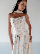 A woman modeling a strapless halter-neck midi dress with a draped bodice and a soft floral print in muted pink, green, and blue tones on a light cream background. / Basque waist, draped bodice A-line gown.