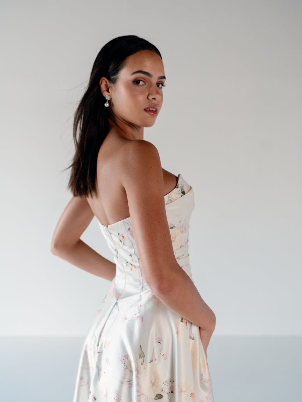 A woman wearing a strapless white satin dress with a delicate pastel floral print, photographed from behind and side against a plain light gray studio background. / Basque waist, draped bodice A-line gown.