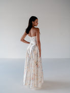 A woman modeling a strapless white satin gown with a floral print, shown from the back against a plain light gray studio background. / Basque waist, draped bodice A-line gown.