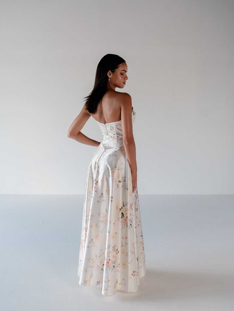 A woman modeling a strapless white satin gown with a floral print, shown from the back against a plain light gray studio background. / Basque waist, draped bodice A-line gown.