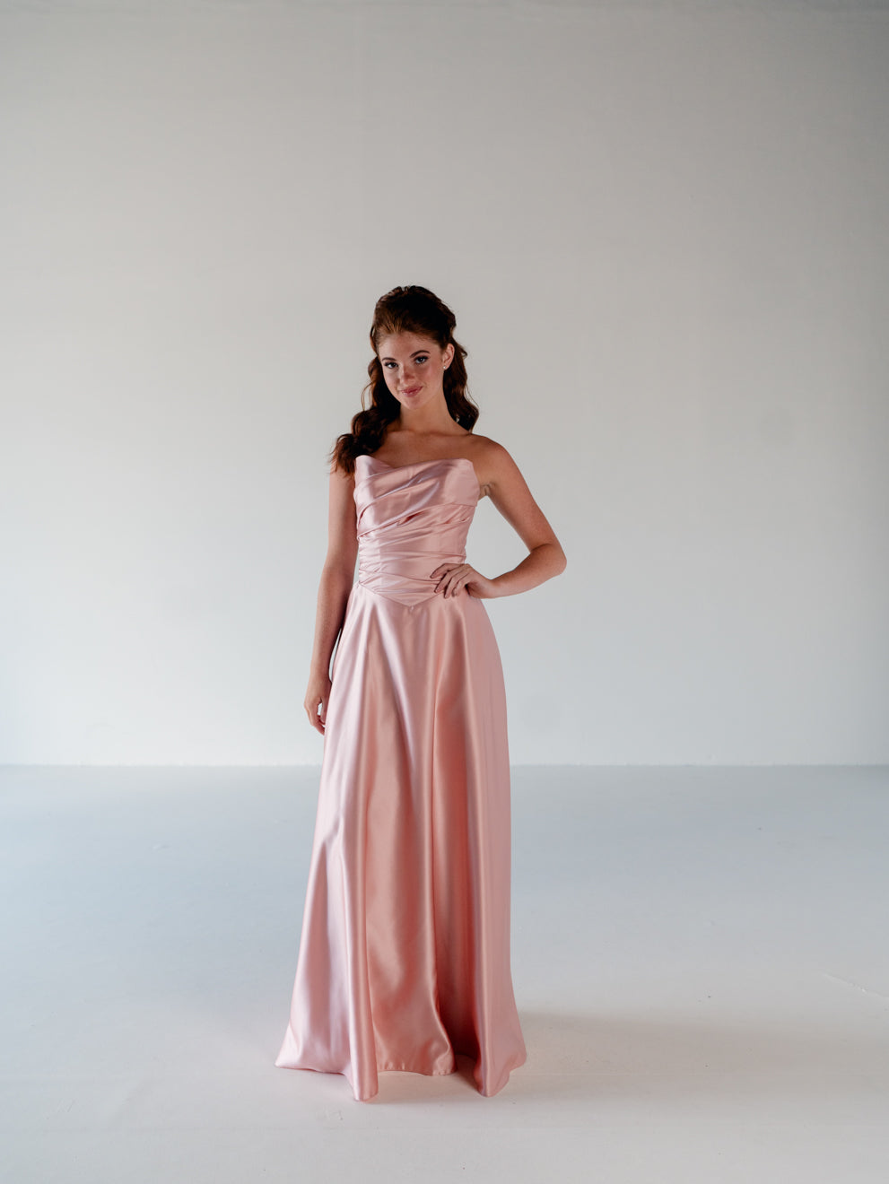 A young woman modeling a strapless, floor-length blush pink satin gown against a plain white studio background. / Basque waist, draped bodice A-line gown.