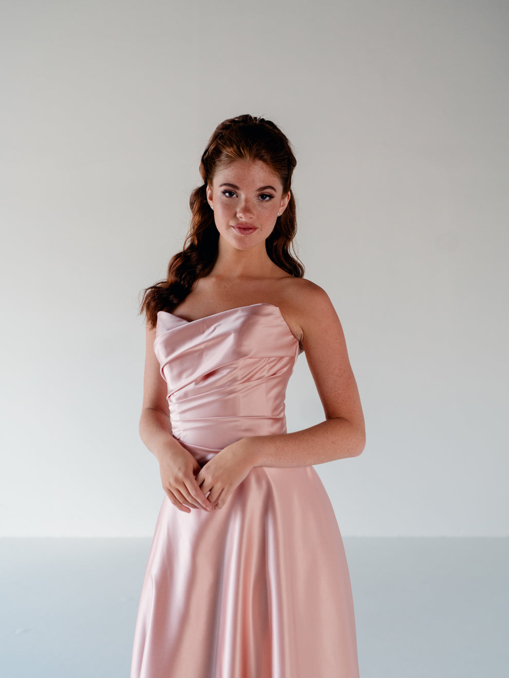 A woman modeling a strapless light pink satin gown with a draped bodice and flowing skirt, standing against a plain light gray studio background. / Basque waist, draped bodice A-line gown.