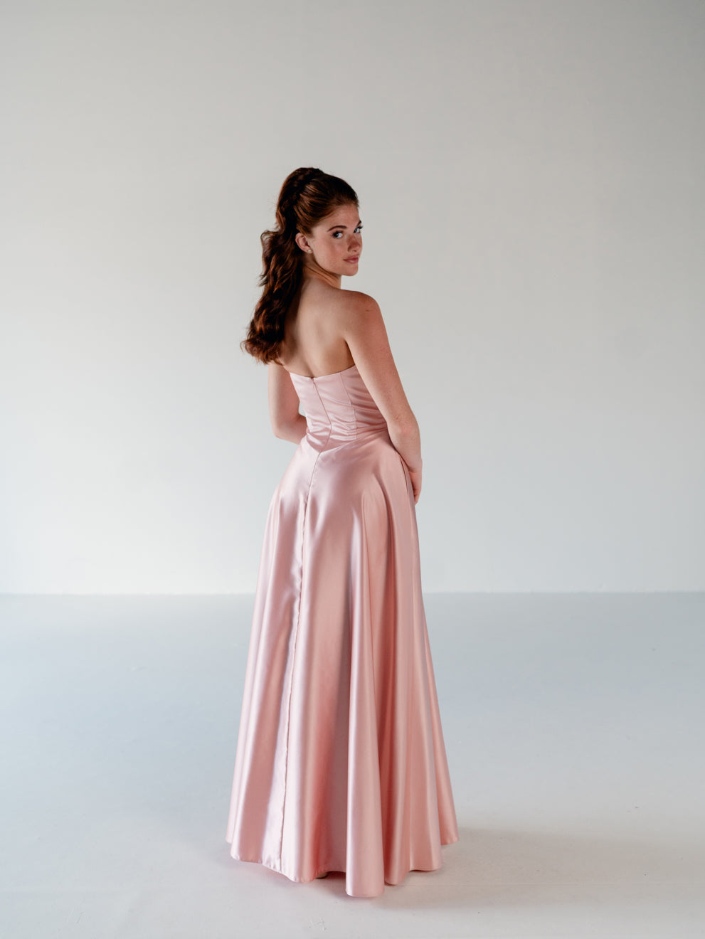 A woman modeling a strapless floor-length gown in a pale blush pink satin-like fabric, photographed in a clean studio setting against a light gray background. / Basque waist, draped bodice A-line gown.